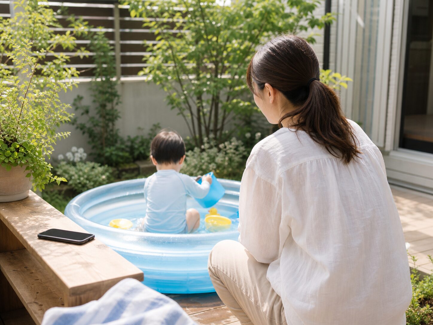 庭の日陰でスマホを置いて子どもの水遊びを見守る親のイメージ